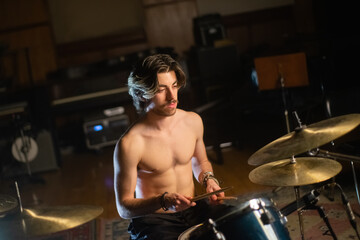 Portrait of young man with moustache playing drum set in garage. Musician sitting shirtless at drum set, training or recording music. Drummer training concept