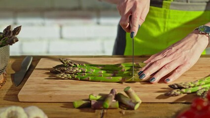 Wall Mural - Woman slicing asparagus and vegetables for cooking on kitchen table. Closeup hands.