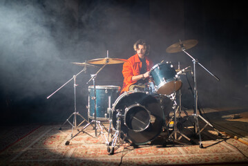 Portrait of young man playing drum set in studio with smoke in background. Musician sitting in spotlight and training or rehearsing music. Drummer training concept