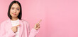 Skeptical asian businesswoman, saleswoman sulking and looking with disapproval while pointing fingers right, showing bad info, upsetting news, standing over pink background