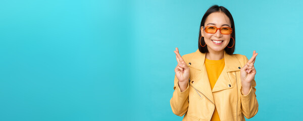 Smiling beautiful asian woman wishing, cross fingers for good luck and looking hopeful, standing over blue background