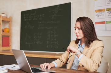 Side view portrait of young female teacher sitting at desk in school classroom and using laptop, copy space