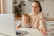 © sementsova321 - Image of puzzled dark haired woman with ponytail wearing beige jumper posing in kitchen and having online conversation, holding toddler daughter in hands, shrugging, being not sure.