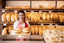Portrait Of Beautiful Smiling Bakery Seller Standing By Shelves Full With Pastries And Bread In Bakery Shop.