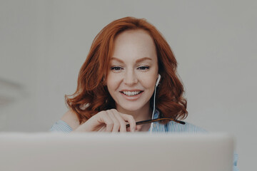 Woman gives online consultation, sits in front of opened laptop computer, has discussion with corporate client, smiles happily, uses modern earphones