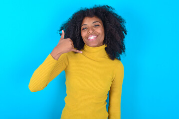 Wall Mural - Young woman with afro hairstyle wearing yellow turtleneck over blue background smiling doing phone gesture with hand and fingers like talking on the telephone. Communicating concepts.