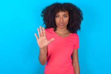 Wall Mural - Young girl with afro hairstyle wearing pink t-shirt over blue background shows stop sign prohibition symbol keeps palm forward to camera with strict expression