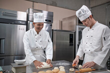 Two Professional Asian Male Chefs In White Cook Uniforms And Aprons Are Kneading Pastry Dough And Eggs, Preparing Bread And Fresh Bakery Food, Baking In Oven At Stainless Steel Kitchen Of Restaurant.
