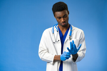 Wall Mural - Young African American doctor putting on surgical gloves on blue background