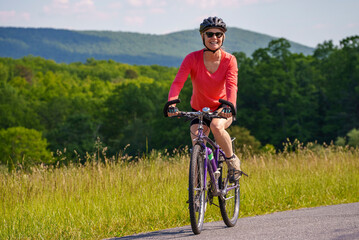 Front view of smiling mature woman biking on a beautiful country road in the mountains on a sunny spring summer day.