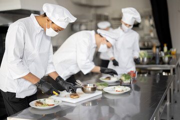 Multiracial group of cooks finishing main courses while working together in the kitchen. Cooks wearing uniform and face mask. Team prepares meals for the restaurant