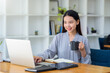 © amnaj - Happy beautiful Asian business woman holding a coffee mug and laptop at the office.