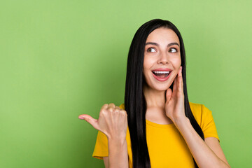 Wall Mural - Photo of excited sweet woman wear yellow t-shirt arm cheek pointing empty space isolated green color background