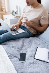 Poster - cropped view of blurred man holding cup of coffee while using laptop in bedroom.
