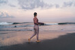 © BullRun - Side view of slim woman in sportive clothes holding mat and walking near calm sea waves spending weekend morning for training at nature, beautiful female in tracksuit choosing place for stretching