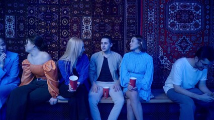 Two girls and a guy are sitting against the background of Soviet carpets and talking in a nightclub under the light of colored spotlights. Night club