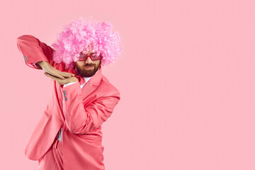 Funny goofy bearded adult man wearing pink party suit, curly clown wig and funky sunglasses dancing isolated on blank empty solid pink background with copy space on right side