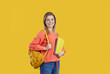 Studio portrait of happy university student with backpack. Pretty young girl in orange sweatshirt and blue jeans standing isolated on yellow background, holding bag and notes and smiling at camera
