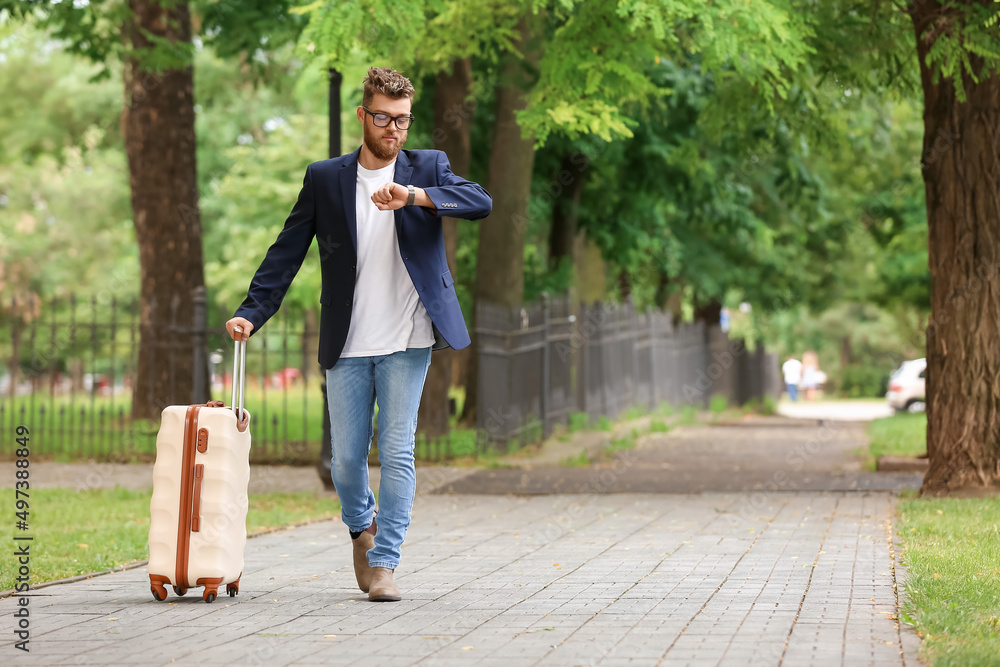 Handsome young man with suitcase looking at wristwatch outdoors