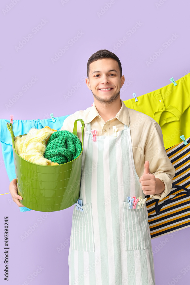 Young man with clothespins and clean laundry showing thumb-up on lilac background
