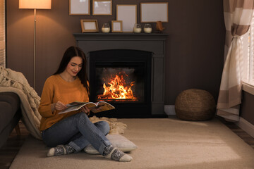 Poster - Beautiful young woman reading magazine on floor near fireplace at home. Space for text