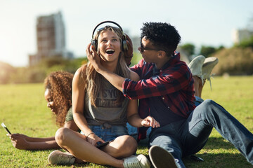 Poster - Have a listen. Shot of a two cheerful young friends listening to music on headphones outside on a park during the day.