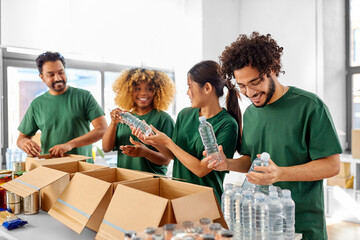 Poster - charity, food donation and volunteering concept - international group of happy smiling volunteers packing bottles of water in boxes at distribution or refugee assistance center