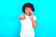 © Jihan - Positive young woman with afro hairstyle wearing white T-shirt against blue wall indicates directly at camera has telephone conversation smiles broadly enjoys talking long hours. You join me