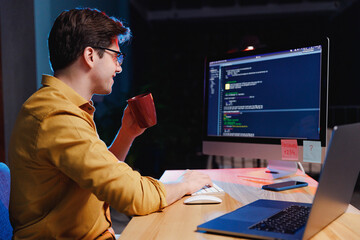 Side profile view young freelancer software engineer IT specialist programmer man in shirt work at night at home writing code on desktop pc computer hold cup drink coffee. Program development concept