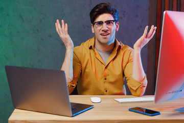 Young questioned sad employee programmer IT specialist business man in casual shirt sit work at office desk with pc laptop desktop computer spread hands Achievement career program development concept.