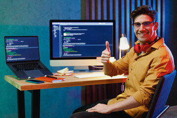 Young fun smiling front-end developer software engineer IT specialist programmer man in shirt work at home writing code on laptop pc destop computer show thumb up gesture. Program development concept.