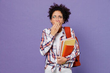 Wall Mural - Young girl woman of African American ethnicity teen student in shirt backpack hold books cover mouth with hand isolated on plain purple background. Education in high school university college concept.