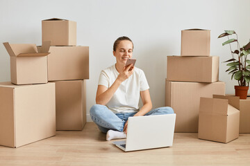 Full length portrait of Caucasian woman wearing white t shirt and jeans sitting on floor surrounded with cardboard boxes and using smart phone for recording voice messages.