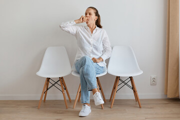 Portrait of sleepy dark haired woman wearing white shirt and jeans sitting on chair in queue, being tire to wait for long hours, yawning, covering mouth with hand.