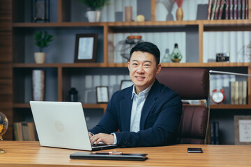 Wall Mural - Portrait of a successful Asian businessman, man working in the office sitting at the table, looking at the camera and smiling, happy banker with laptop