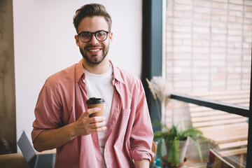 Wall Mural - Half length portrait of cheerful hipster guy in optical spectacles for provide eyes protection smiling at camera during coffee break in coworking space or cafe interior, happy young man with paper cup