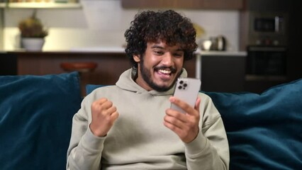 Happy satisfied Indian man holding smartphone and smiling making yes gesture, multiracial guy celebrating online lottery or giveaway victory sitting on the sofa at home