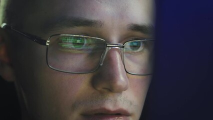 The programmer in glasses looks at the monitor screen. He's engrossed. Close-up of his face. The glasses reflect the lines of code for the program he creates
