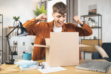 Wall Mural - Handsome young man gesturing while unpacking parcel, excited, clenches fists in triumph. Joyful male vlogger. Concept of people and social networks.