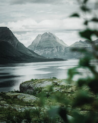 Wall Mural - Beautiful view of mossy rocks by a shiny lake under snow mountains against a cloudy gray sky