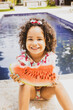 © EZ PHOTOS - Portrait of smiling baby girl with afro hair style  in a colourful swimsuit outfit eating watermelon by the pool. Afro-American and Afro-Brazilian children summer concept with copy space.