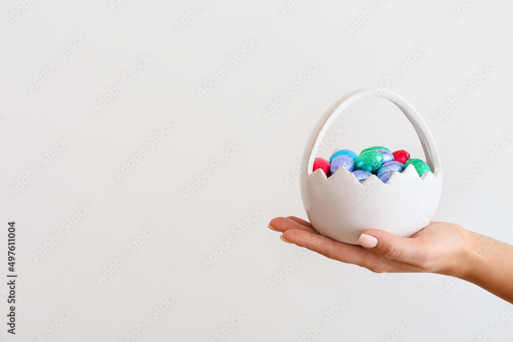 Female hand with ceramic basket of chocolate eggs on light background