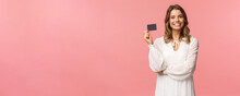 Portrait Of Pleased Good-looking Blond European Female In White Dress, Show Credit Card With Satisfied Expression, Smiling Camera, Recommend Bank Services, Use Payment Online, Pink Background