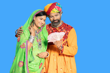 Happy indian couple wearing traditional colorful outfit holding indian rupees note against blue background, husband and with with lots of money in hand, savings and financial growth.