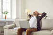 Smiling man puts his hands behind his head taking break after working with laptop. Man enjoys productive work sitting on comfortable sofa in living room. Leisure and business online concept.