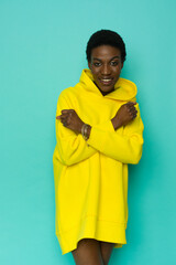 Wall Mural - Happy young black woman in vibrant yellow oversized hooded sweatshirt is posing with arms crossed.