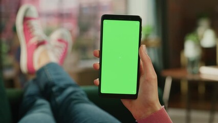 Fototapete - Feminine Hand Holding a Smartphone with Green Screen Mock Up Display. Female is Relaxing on a Couch at Home, Watching Videos and Reading Social Media Posts on Mobile Device. Close Up POV Footage.
