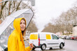 Horizontal front view of woman crossing the street with umbrella outdoors. Mid waist view of woman in yellow raincoat with van driving in the background. Weather concept