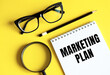 © Nastassia - The phrase MARKETING PLAN in notebook with pencil, magnifier, and glasses on a yellow background