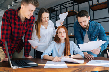 Tired young female team leader under stress at office sitting at desk with multiracial colleagues shaking paper documents standing near her, looking at camera. Woman boss exhausted by management work.